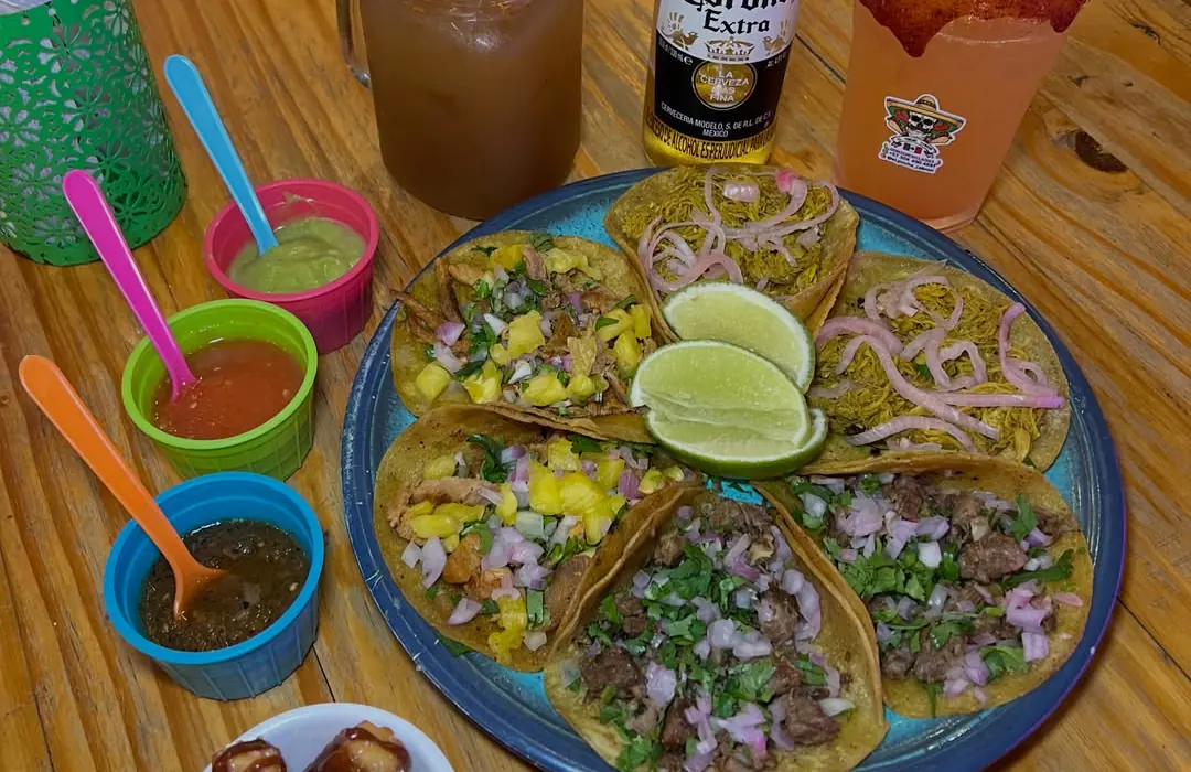 Plato de tacos con limones, salsas de colores, agua fresca y una cerveza sobre mesa de madera